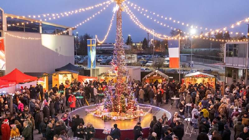 Eine festliche Weihnachtsmarktkulisse zeigt einen hohen, beleuchteten Baum, der mit Lichtern und Dekorationen geschmückt ist. Menschenmengen in Winterkleidung versammeln sich um festlich geschmückte Stände und genießen die lebhafte, fröhliche Atmosphäre.