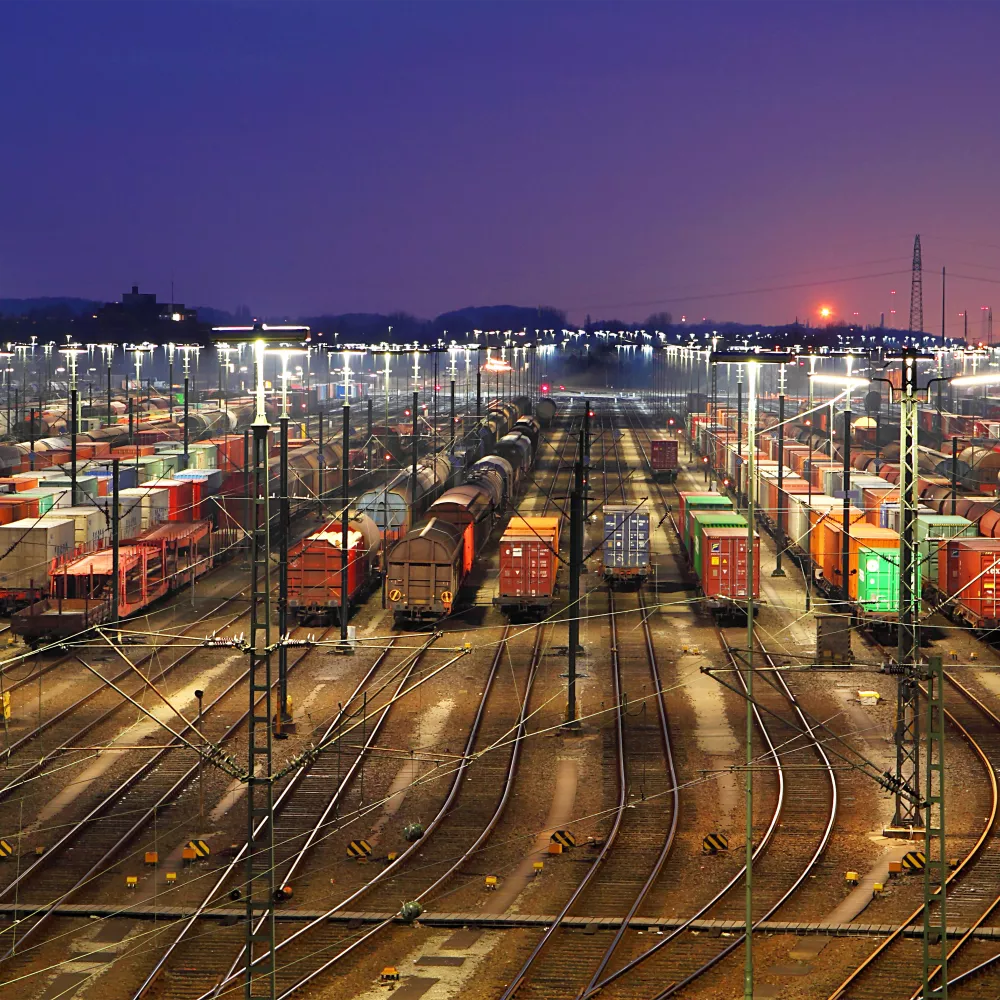 Zahlreiche Güterzüge und Container stehen in der Abenddämmerung in einem hell erleuchteten Rangierbahnhof, während die Gleise in der Ferne zusammenlaufen und die Sonne am Horizont untergeht.