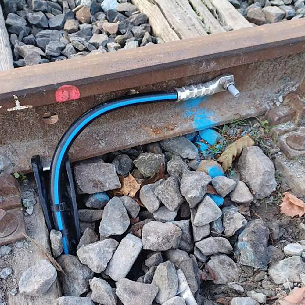 A black and blue cable with a metal fitting runs under railway tracks, laying among rocks and next to wooden sleepers, with blue paint marking on the rail and stones.