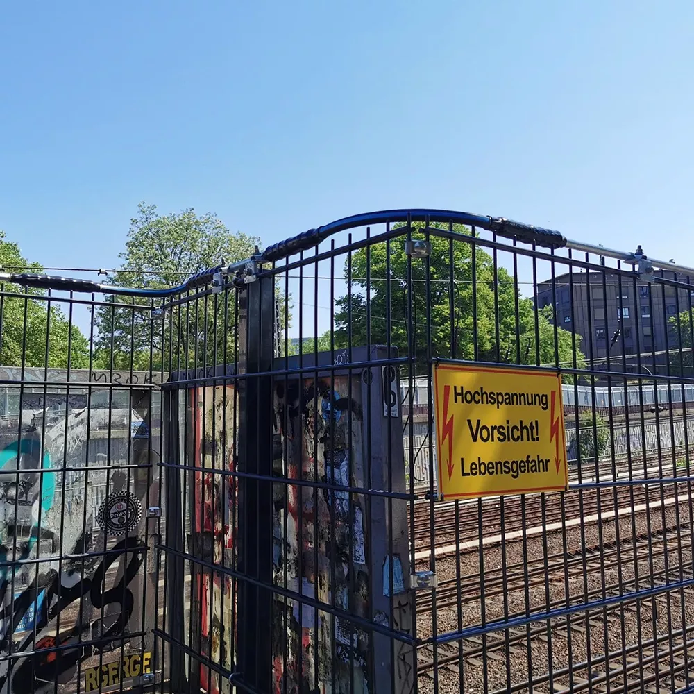 Fenced railway area with a yellow high-voltage warning sign under a clear blue sky.